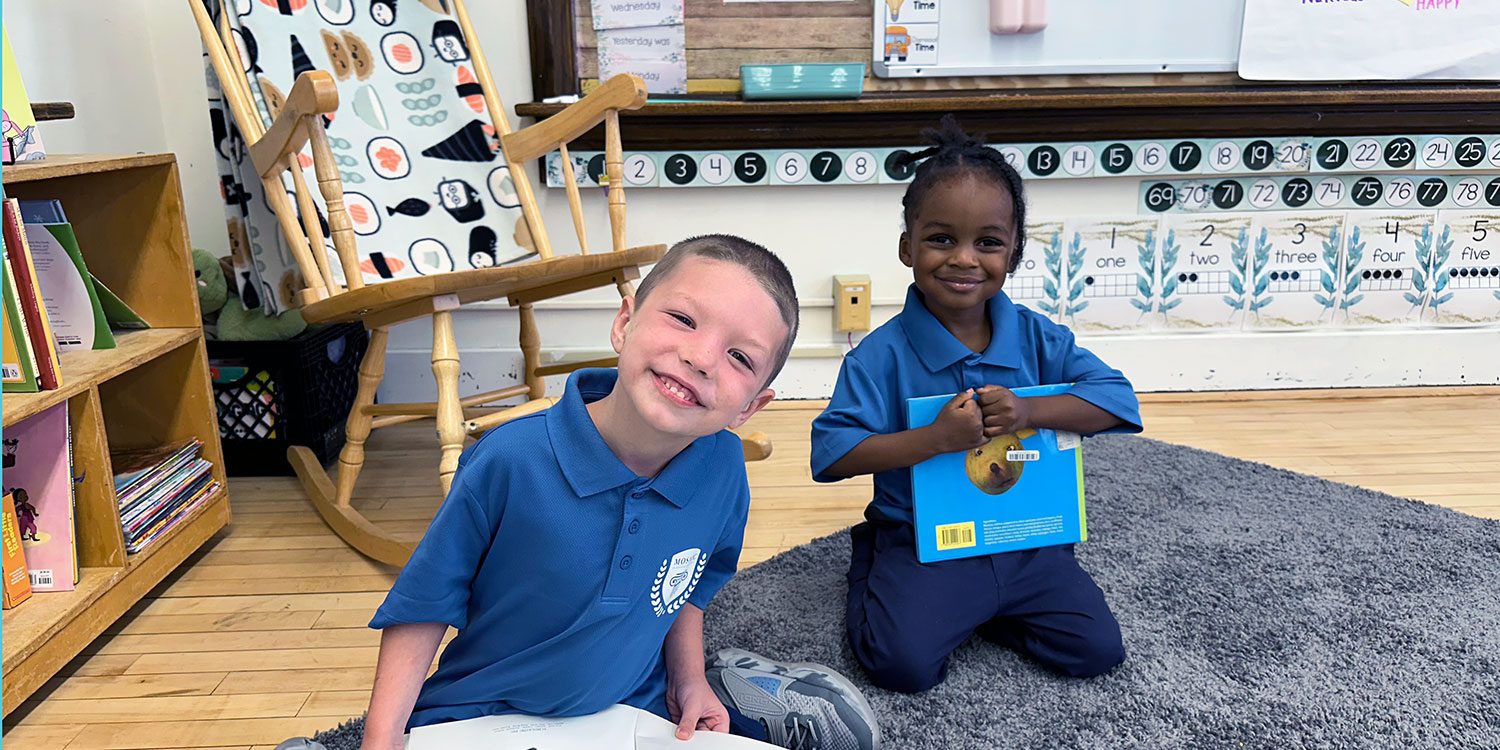 Two students sitting on a classroom rug, one holding a book.