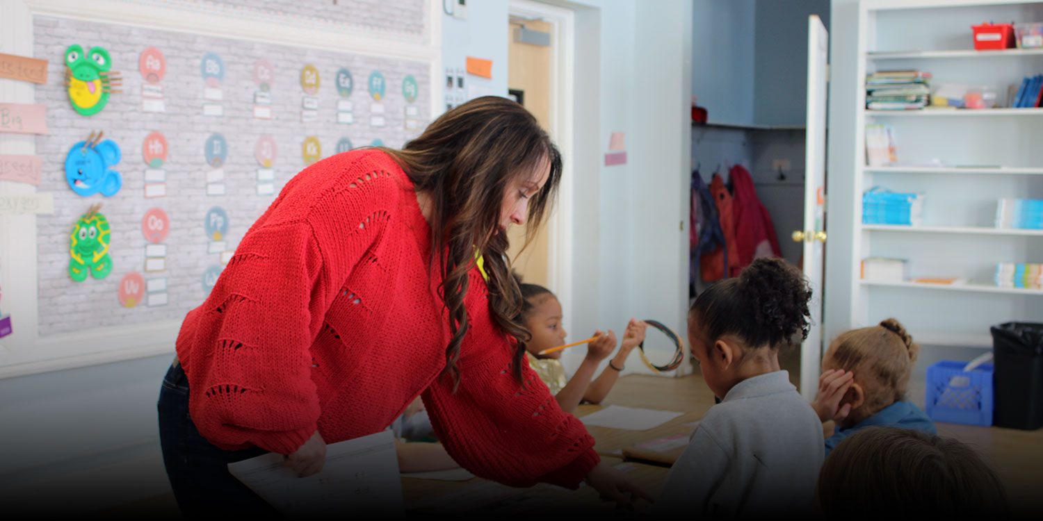 Teacher assisting students and pointing to a worksheet for a student while they are working at a desk